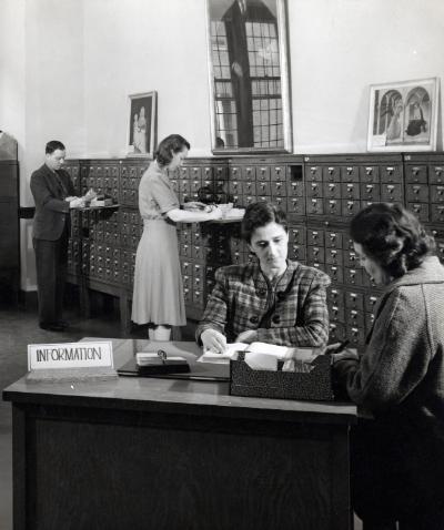 Historical photograph of a librarian at an information desk consulting with a student and two people behind using the card catalog