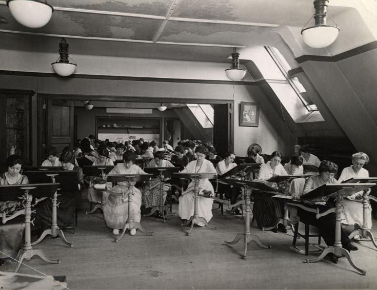 Black and white photograph depicting a group of women sitting at small desks. They each have pencils and paper and are sketching.
