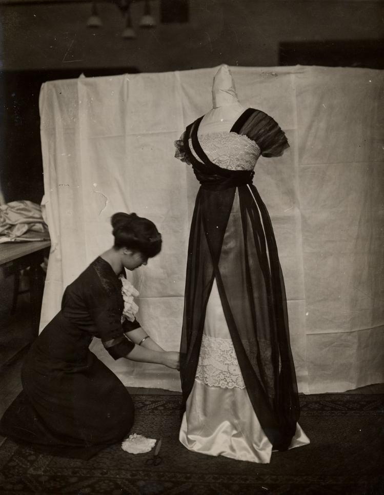 Student (lower left) dressed in dark 1910s style clothing, draping a dress model with a new dress design (right).