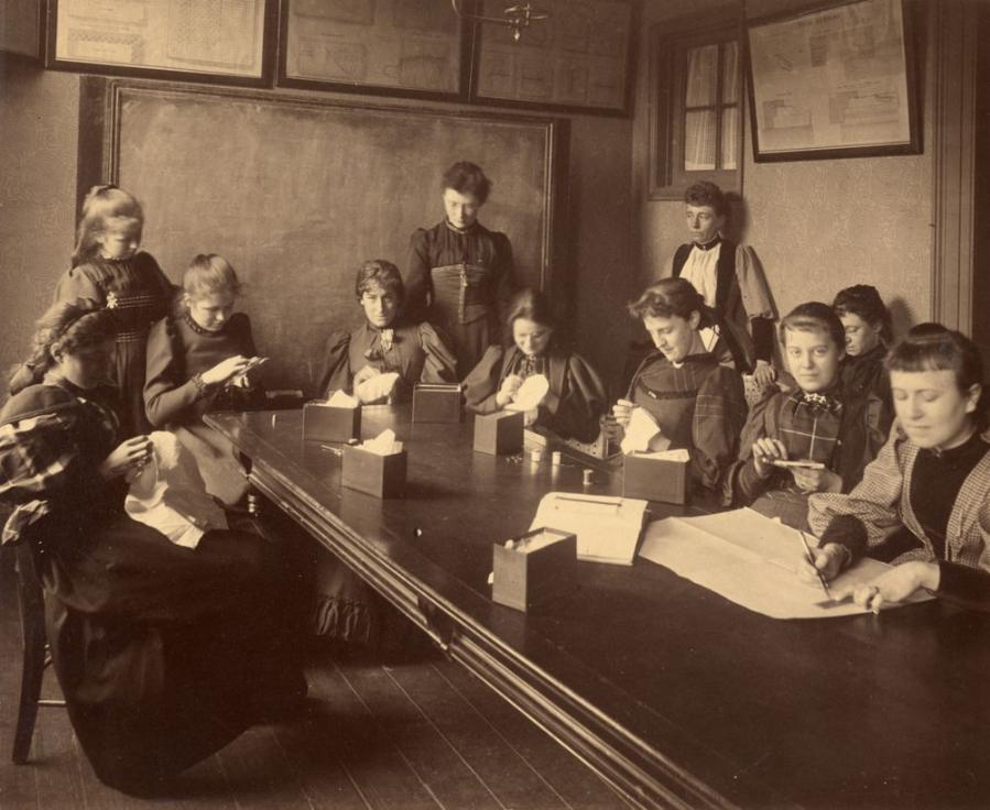 A group of 11 young women sit around a long, rectangular table. The women are engaged in various sewing exercises and are dressed in late 1890s clothing.
