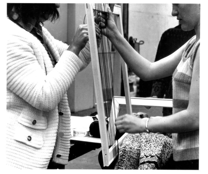 Two women tapestry weaving. One stands on the left and the other stands on the right of the loom's frame.