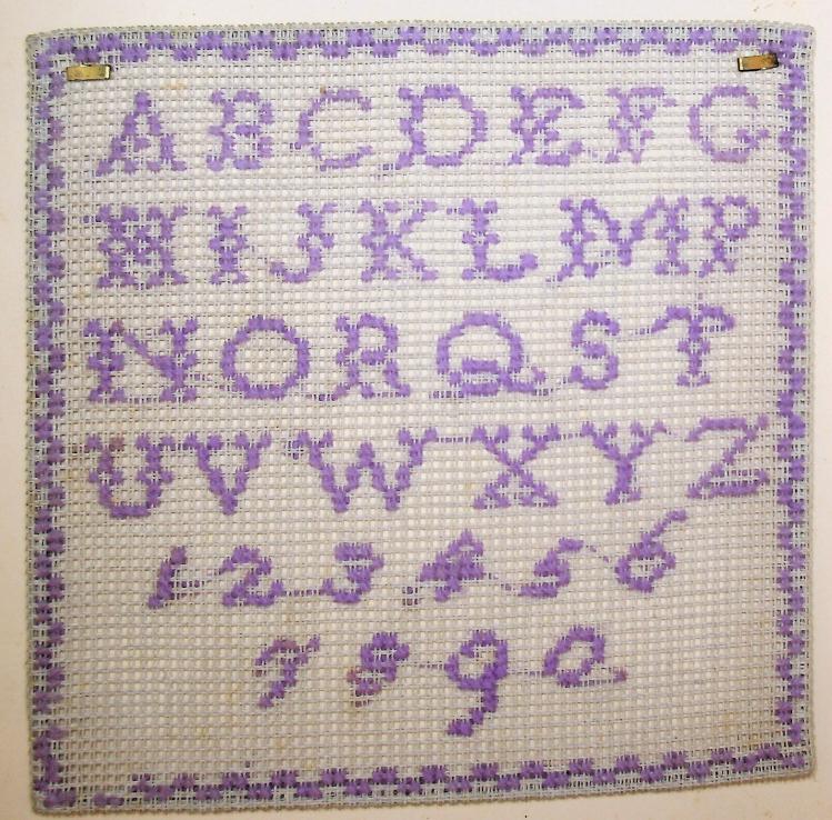 An example of a sewing sampler. The alphabet and the first 9 numerals are stitched in purple thread onto white cloth